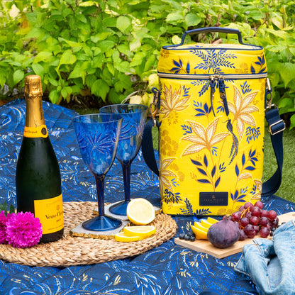 Yellow floral-patterned cooler bag with wine bottle, glasses, and fruits on a blue tablecloth outdoors.