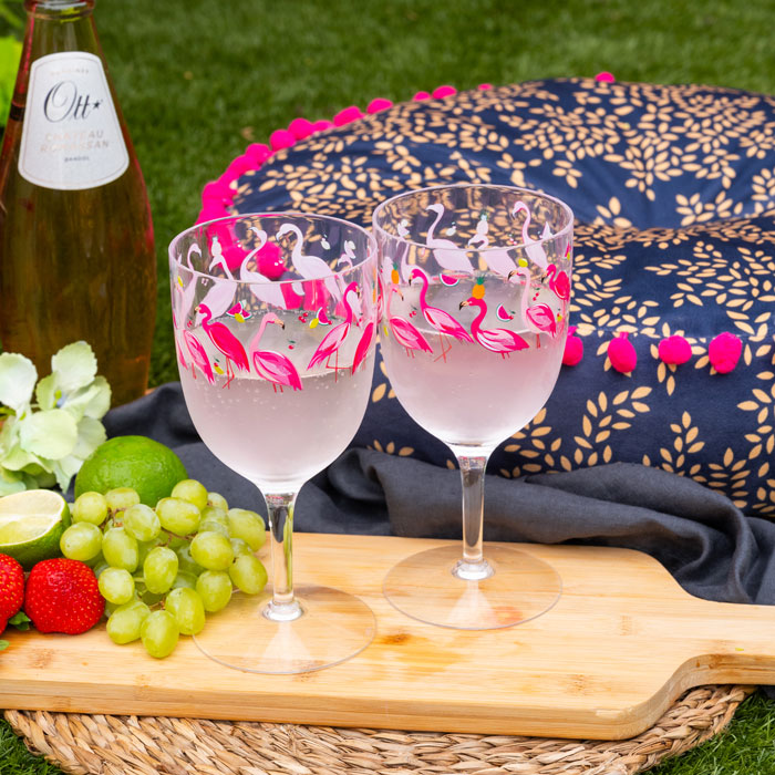 Two plastic wine glasses with flamingo design, bottle of wine, fruits, and a blanket on a grassy background.