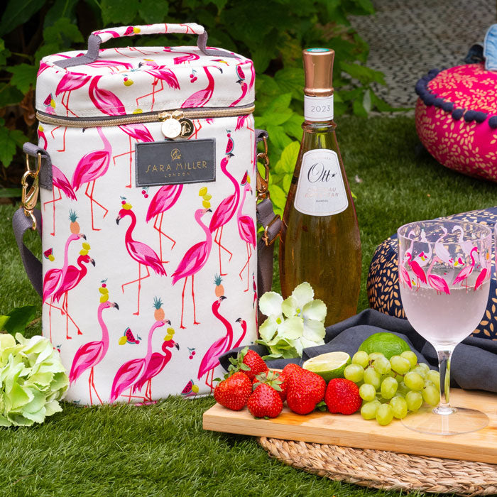 Flamingo patterned cool bag with a bottle of wine, fruits, and a glass on a grassy background.