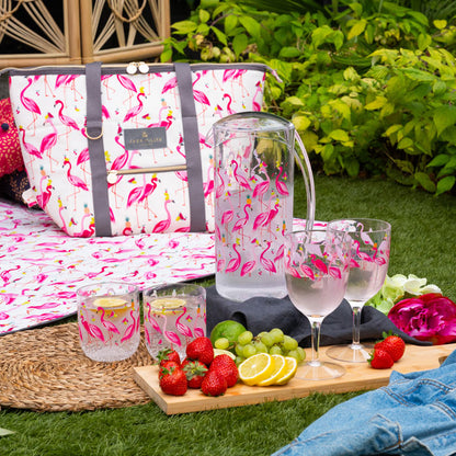 Picnic setup with flamingo-patterned pitcher, plastic cups, and food on a grassy area.