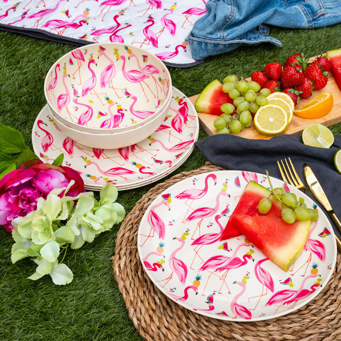 Flamingo-patterned plastic plates and bowls on a picnic setup with fruit and flowers.
