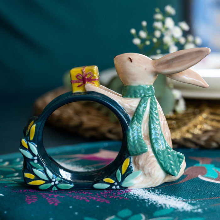 A ceramic green napkin ring featuring a rabbit wearing a green scarf and holding a small yellow gift box with green and yellow foliage accents.
