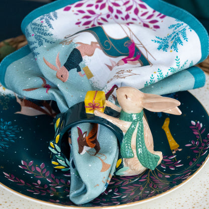 The napkin with woodland animals playing instruments styled inside a matching ceramic napkin ring, at a table setting.