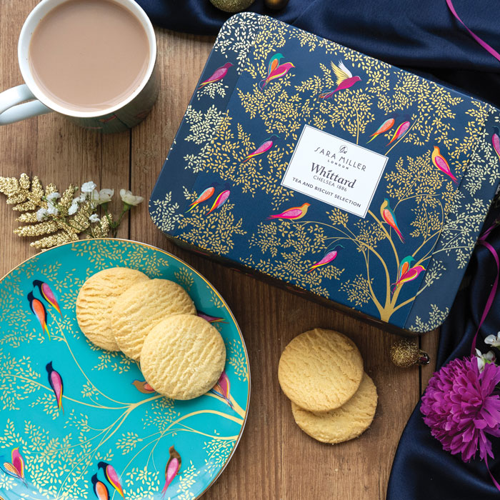 Tea and biscuit selection tin with Smokey Birds design on a table, with mug of tea and plate with shortbreads.