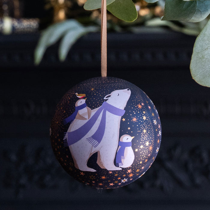 A spherical Christmas ornament with a dark navy blue background, decorated with illustrations of a polar bear, penguin, and a small robin wearing purple scarves, hanging by a thin brown ribbon.