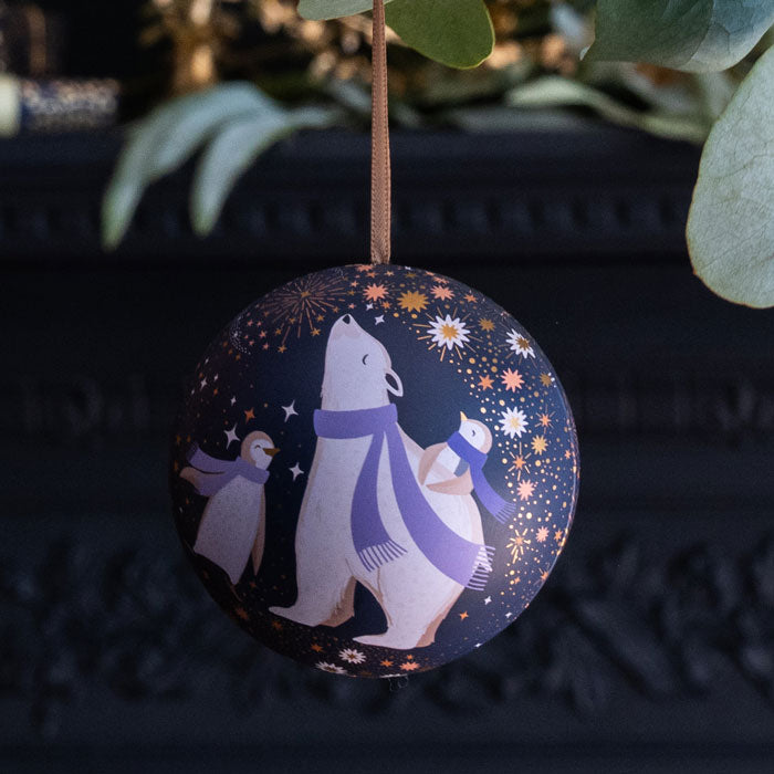 A round hanging ornament featuring a whimsical winter scene with a polar bear, penguin, and small robin under a starry night sky, suspended by a brown ribbon.