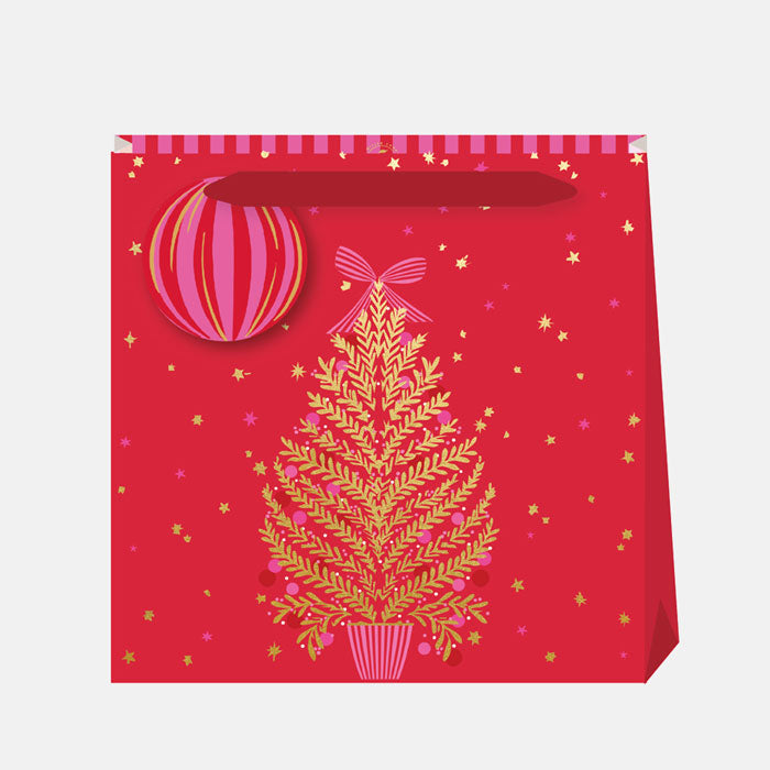 A Christmas gift bag with a gold tree and pink decorations and bow, with gold stars and a red background, matching gift tag and  ribbon handles.
