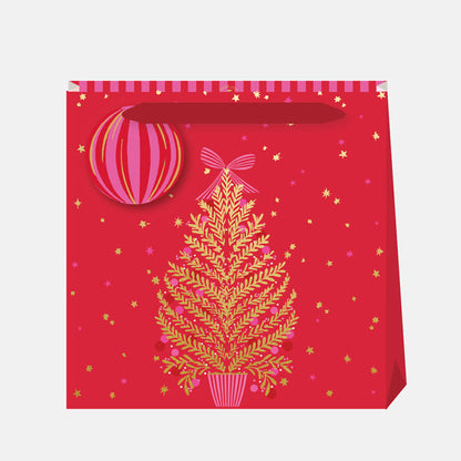 A Christmas gift bag with a gold tree and pink decorations and bow, with gold stars and a red background, matching gift tag and  ribbon handles.
