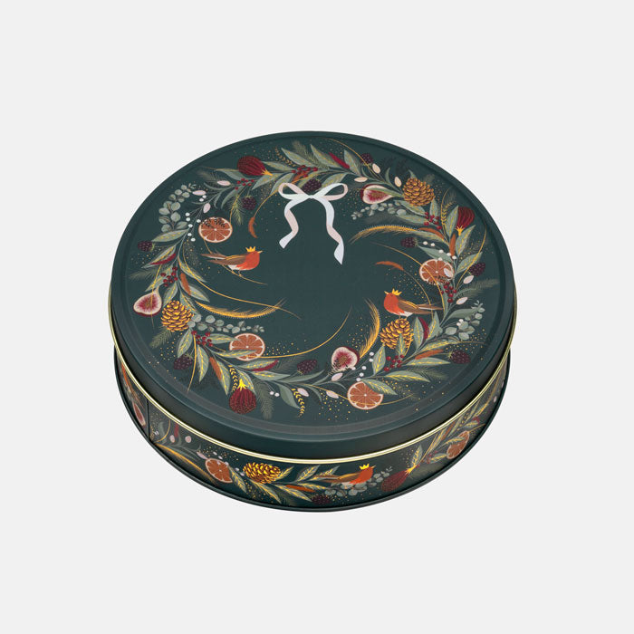 A round decorative tin with a dark green lid featuring a hand-painted wreath of leaves, berries, small birds, and citrus slices, accented with a white bow motif at the top center.