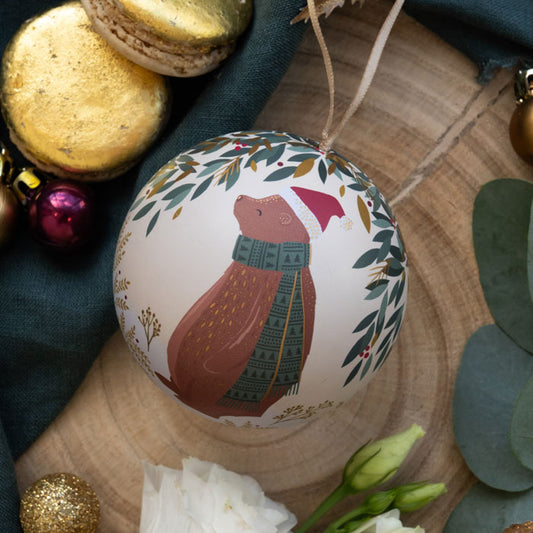 A round Christmas ornament featuring a  brown bear wearing a red Santa hat and green patterned scarf, surrounded by festive foliage details on a cream background. 