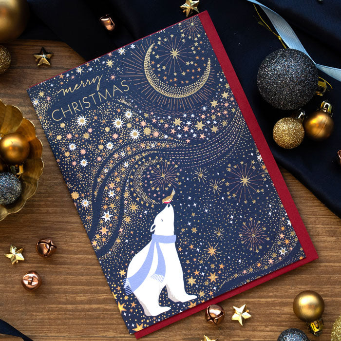 A Christmas greeting card with a navy blue background, gold stars and crescent moon, and a white polar bear wearing a blue scarf, styled on a wooden surface with holiday ornaments.
