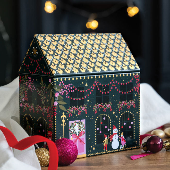 A decorative house-shaped tin with a dark green and gold patterned roof and festive holiday illustrations on the front, styled on a tabletop with ornaments, a red ribbon, and blurred string lights in the background.
