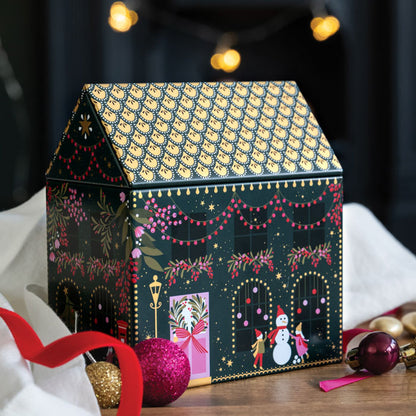 A decorative house-shaped tin with a dark green and gold patterned roof and festive holiday illustrations on the front, styled on a tabletop with ornaments, a red ribbon, and blurred string lights in the background.