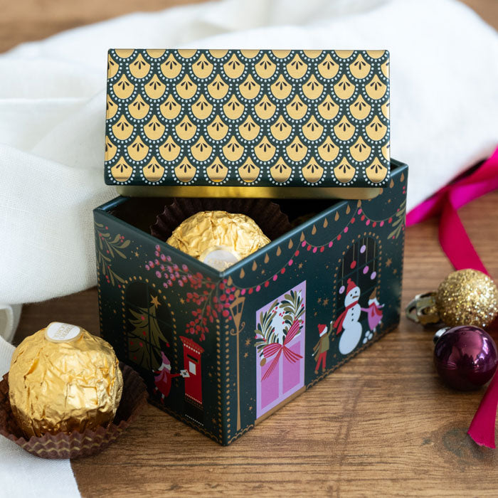 A decorative house shaped tin decorated with a festive facade filled with Ferrero Rocher chocolates, styled on a wooden surface with holiday ornaments and a white fabric background.