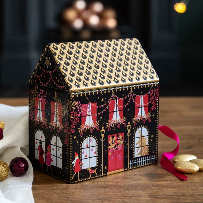 A decorative house-shaped tin with a black and gold patterned roof and facade, decorated with red curtains, pink garlands, and holiday figures, set on a wooden surface with festive ornaments nearby.