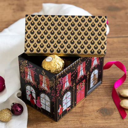 A decorative tin with a black and gold patterned lid and a house-themed printed exterior, shown open on a wooden surface with a Ferrero Rocher chocolate inside and festive props nearby.