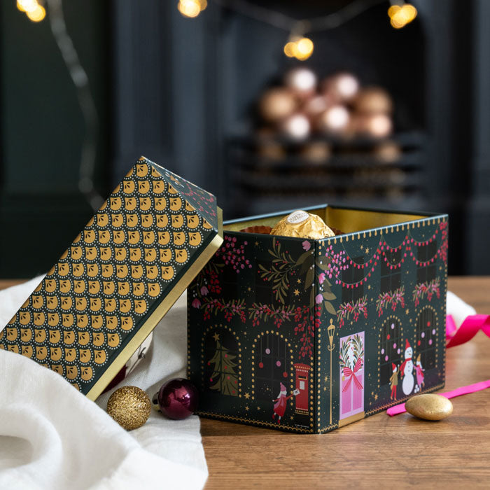 A decorative black Christmas-themed tin  filled with Ferrero Rocher chocolates, set on a wooden table with holiday ornaments and warm blurred lights in the background.