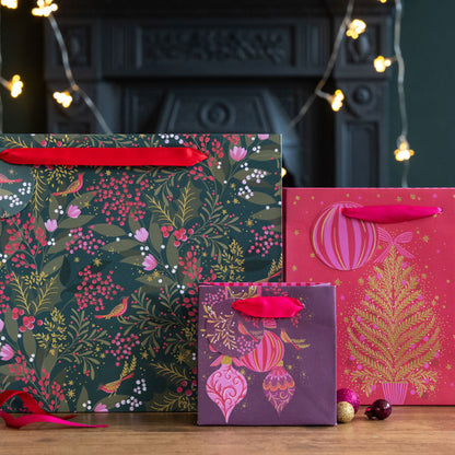 Three Christmas-themed gift bags with red ribbon handles. The large bag features a dark green festive foliage pattern with birds and berries, the right bag is bright pink with a gold Christmas tree, and the small bag is purple with pink ornaments.
