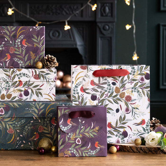 A set of three gift boxes with a fig and fern design on them in greens and reds with little crowned robins, one in plum one in green and one in cream, next to matching gift bags.
