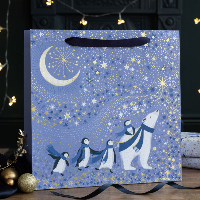 A periwinkle gift bag with little penguins surrounding a polar bear under a crescent moon and starry sky, with gold embellishment.