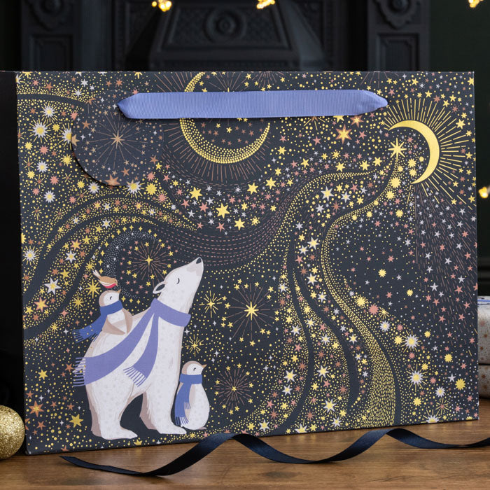 A shopper style gift bag with polar bear and penguins dressed in purple scarves stargazing at the sky and crescent moon, on a navy background with gold embellishment