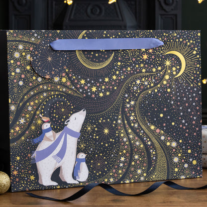 A shopper style gift bag with polar bear and penguins dressed in purple scarves stargazing at the sky and crescent moon, on a navy background with gold embellishment