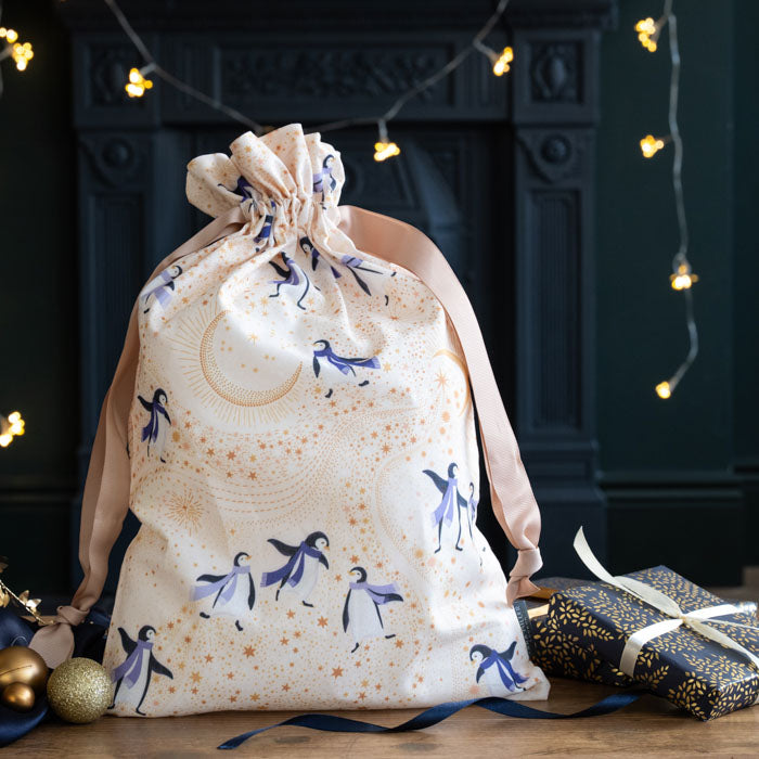 A cream-coloured fabric gift bag with a drawstring closure, decorated with a celestial pattern of penguins, stars, and moons in navy blue and gold. T
