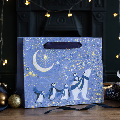 Periwinkle festive gift bag with a polar bear surrounded by penguins all wearing purple scarves, under a starry sky and crescent moon with gold embellishment.