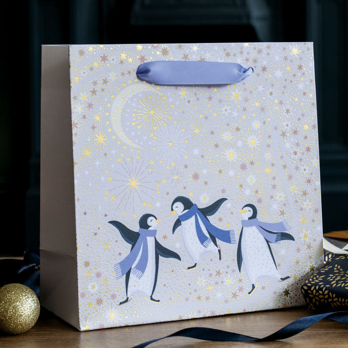 A cream festive gift bag with three penguins dancing wearing purple scarves, under a starry sky and crescent moon with gold embellishment.