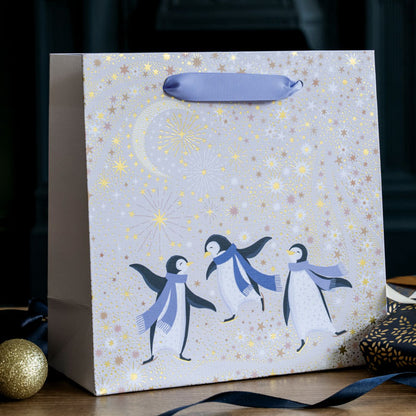 A cream festive gift bag with three penguins dancing wearing purple scarves, under a starry sky and crescent moon with gold embellishment.