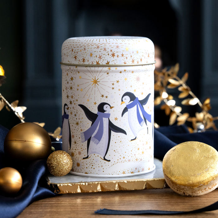 A cylindrical tin canister with a cream lid and body, decorated with three penguins wearing blue scarves and gold stars, styled on a wooden surface with gold ornaments and a macaron.