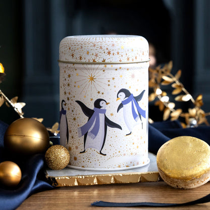A cylindrical tin canister with a cream lid and body, decorated with three penguins wearing blue scarves and gold stars, styled on a wooden surface with gold ornaments and a macaron.