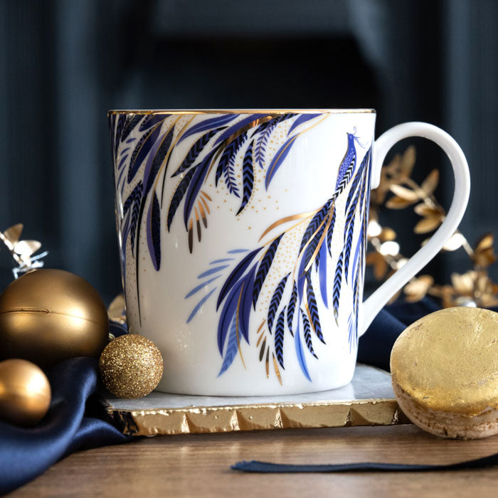 A white and indigo mug with peacock tail feathers design, with gold embellishment surrounded by Christmas accents.