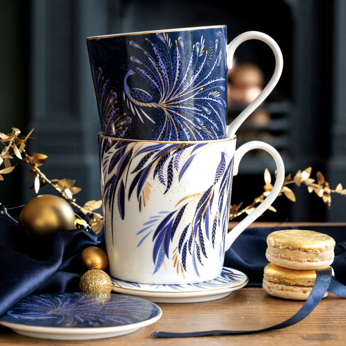 Two mugs stacked, one indigo one white both with gold embellishment with their matching coasters surrounded by Christmas accents.