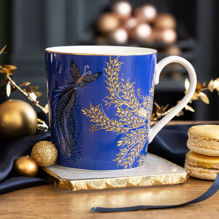 A cobalt mug with butterflies and foliage, gold embellishment, sat on a marble coaster and surrounded by Christmas accents.