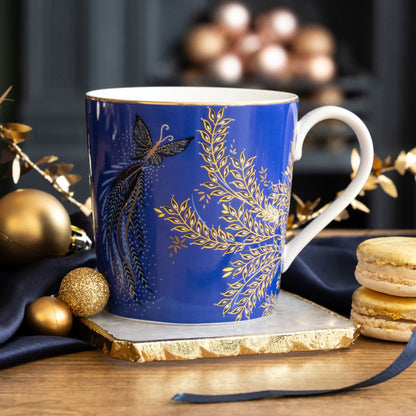 A cobalt mug with butterflies and foliage, gold embellishment, sat on a marble coaster and surrounded by Christmas accents.