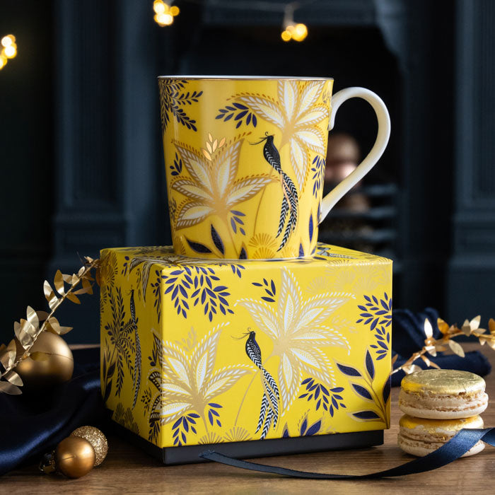 A yellow mug with botanic print, gold embellishment, sat on top of its matching gift box, surrounded by Christmas accents.