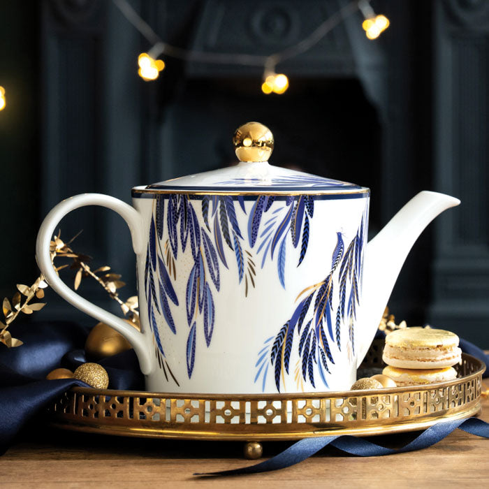 White teapot with indigo peacock tail feather design, gold embellishment, surrounded by Christmas accents.