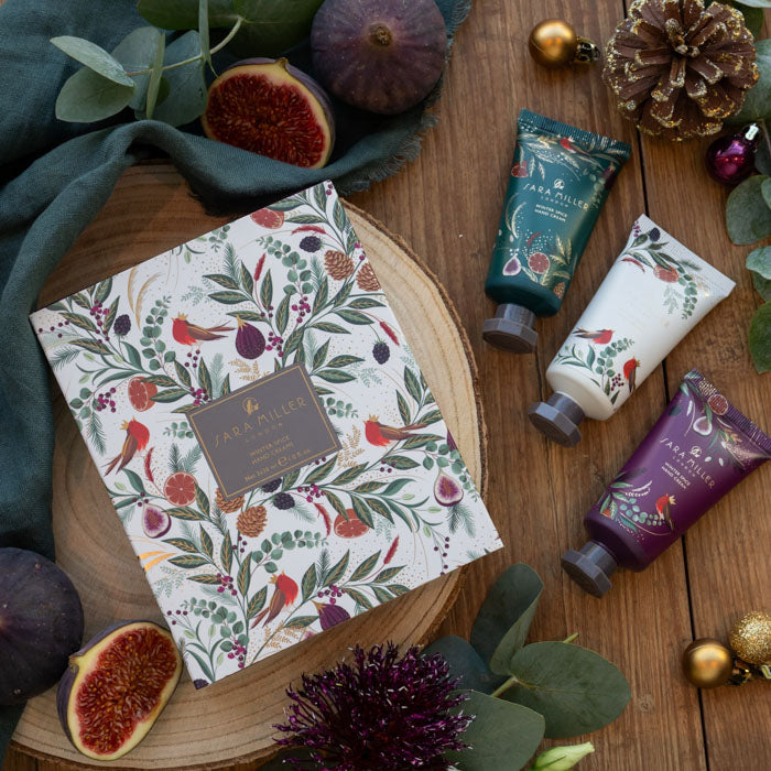 A flat-lay product arrangement featuring a boxed set and three hand cream tubes from Sara Miller London. The products are decorated with a botanical and robin pattern.