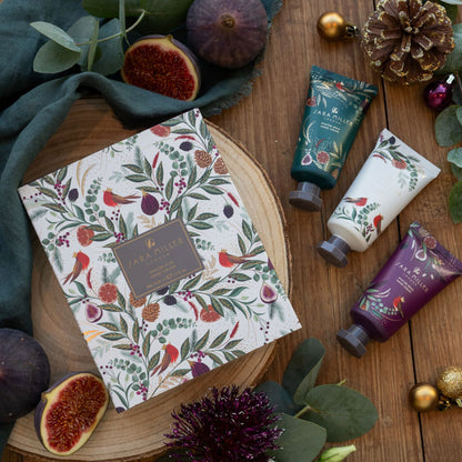 A flat-lay product arrangement featuring a boxed set and three hand cream tubes from Sara Miller London. The products are decorated with a botanical and robin pattern.