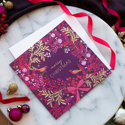 A festive Christmas greeting card with a rich plum and purple background, decorated with gold botanical patterns, red berries, pink birds, and the words 'Merry Christmas' in gold. The card is displayed on a white marble tray with a white envelope, red ribbons, and holiday ornaments.