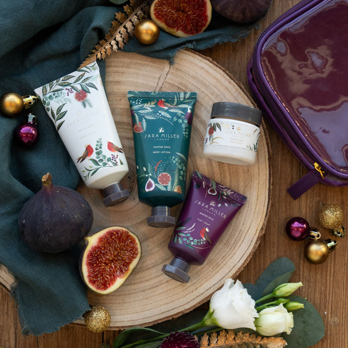 Four Sara Miller body care products arranged on a round wood slice with figs, flowers, ornaments, and a purple zip case on a wooden surface.