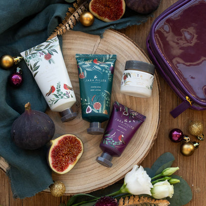 Four Sara Miller body care products arranged on a round wood slice with figs, flowers, ornaments, and a purple zip case on a wooden surface.