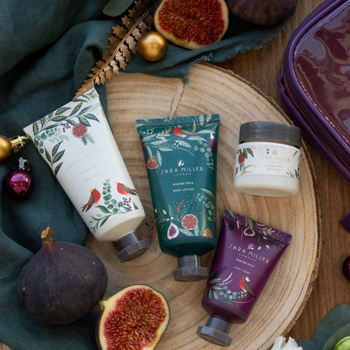 Four Sara Miller body care products arranged on a round wood slice with figs, dark green fabric, and holiday ornaments. The set includes a body wash, body lotion, body butter, and a jar, all decorated with botanical and bird illustrations in white, teal, and purple tones.