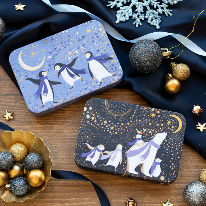 Two decorative rectangular tins decorated with penguins and polar bears dressed in blue scarves. The top box shows penguins under a crescent moon in a light blue starry sky; the bottom box shows a polar bear and penguins under a crescent moon in a dark navy starry sky.
