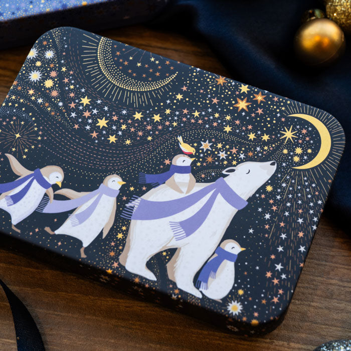 A rectangular tin with rounded corners featuring a whimsical winter night scene. The tin is dark navy blue with gold stars, crescent moons, and shows three penguins and a polar bear wearing matching lavender-blue scarves.