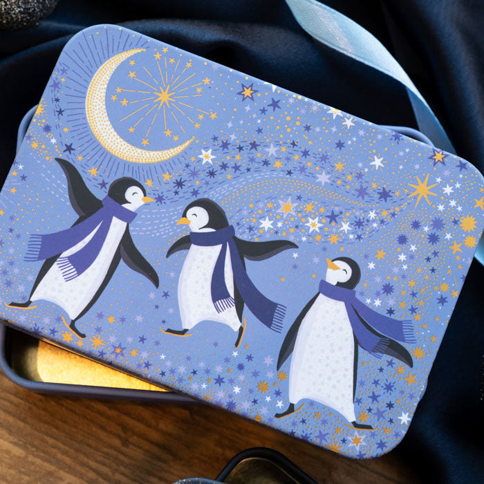 A rectangular tin with rounded corners, featuring a blue penguin-themed design with three penguins wearing purple scarves, a crescent moon, and stars on a periwinkle background.