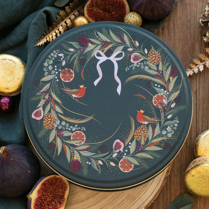A round decorative tin with a festive wreath design, featuring red robins, pinecones, citrus slices, and a white bow motif on a dark green background.