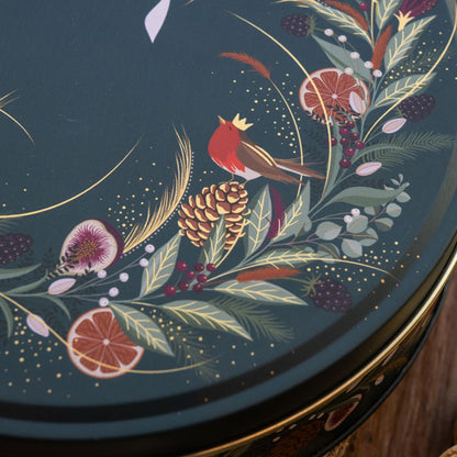 Close-up of a round decorative tin lid featuring a dark green background with hand-painted botanical and bird motifs, including a red robin, pinecone, citrus slices, berries, and gold leaf accents.