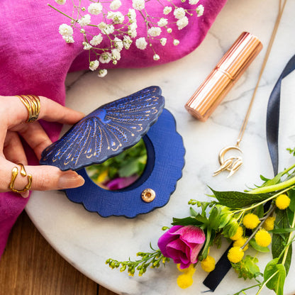 Compact mirror shaped like a peacock with blue and gold peacock design. Hand holding open, can see press stud closure.
